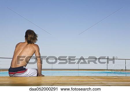 Stock Image - Man sitting poolside, dipping feet in water, rear view. Fotosearch