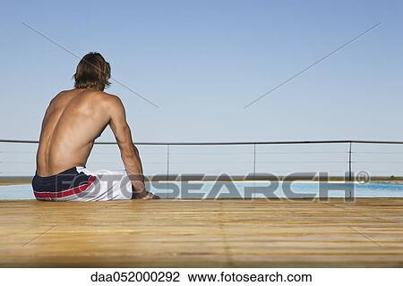 Stock Image - Man sitting poolside, dipping feet in water, rear view. Fotosearch
