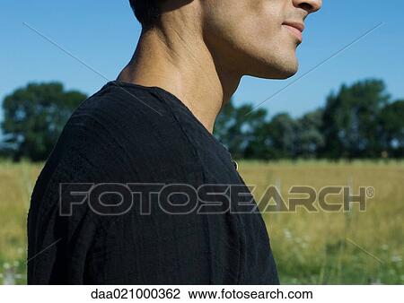 Stock Image - Man standing by field, partial view of shoulders and lower face, side view. Fotosearch