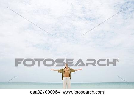 Picture - Man standing in front of sea with head back, shirt open, and arms out, mid-distance. Fotosearch