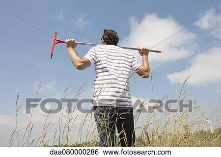 Man standing in grass with rake on shoulders, rear view View Large Photo Image Stock Photograph - Man standing in grass with rake on shoulders, rear view. Fotosearch