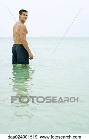 Man standing knee deep in ocean, smiling over shoulder at camera View Large Photo Image Stock Photo - Man standing knee deep in ocean, smiling over shoulder at camera. Fotosearch