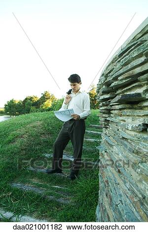 Stock Image - Man standing near stone wall, holding blueprints. Fotosearch