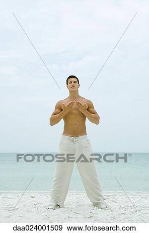 Stock Photo - Man standing on beach, doing yoga, full length. Fotosearch