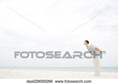 Stock Photograph - Man standing on beach, leaning into wind, arms behind back and eyes closed. Fotosearch