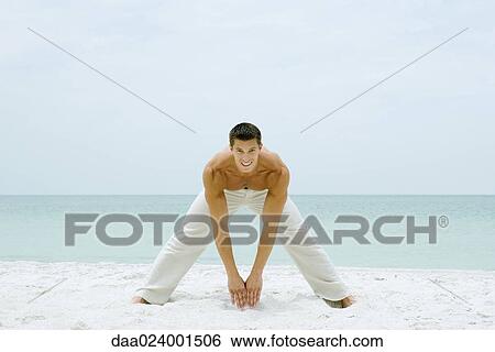 Stock Photograph - Man standing on beach, stretching and smiling at camera, full length. Fotosearch