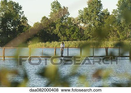 Man standing on footbridge over river in distance, seen through leaves in blurred foreground View Large Photo Image Stock Photo - Man standing on footbridge over river in distance, seen through leaves in blurred foreground. Fotosearch