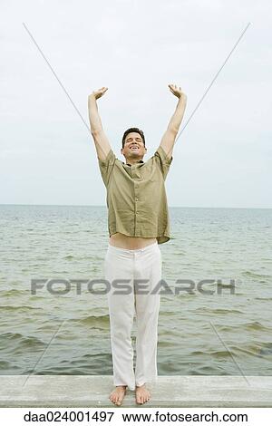 Stock Photo - Man stretching, ocean in background, full length. Fotosearch