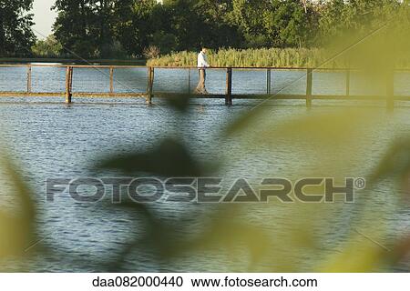 Stock Image - Man walking on footbridge over river in distance, seen through leaves in blurred foreground. Fotosearch