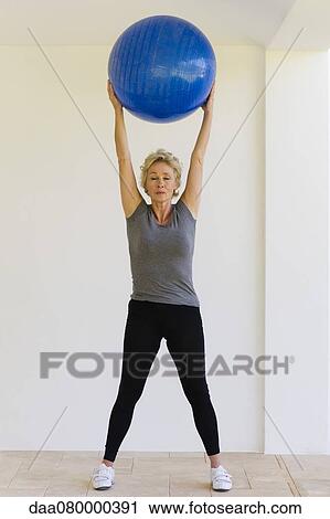 Stock Image - Mature woman holding fitness ball overhead with eyes closed. Fotosearch
