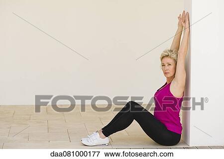 Stock Photo - Mature woman sitting against wall, stretching arms. Fotosearch