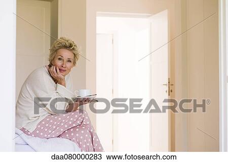 Mature woman sitting on edge of bed with cup of coffee View Large Photo Image Stock Image - Mature woman sitting on edge of bed with cup of coffee. Fotosearch