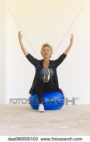Stock Image - Mature woman sitting on fitness ball with arms raised and one leg up. Fotosearch