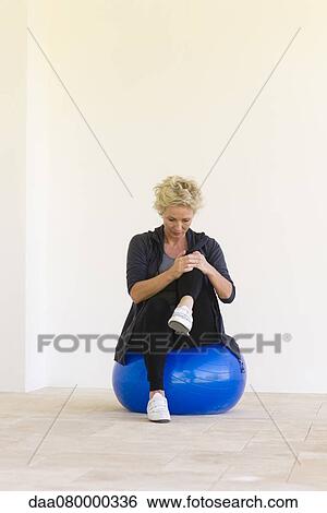 Stock Photograph - Mature woman sitting on fitness ball, hugging one knee. Fotosearch