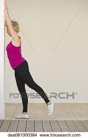 Picture - Mature woman stretching arms against wall while standing on one leg, side view. Fotosearch