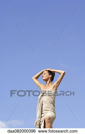 Stock Photograph - Mdi-adult woman standing wrapped in towel against blue sky, hands on head. Fotosearch
