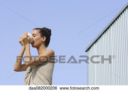 Stock Photograph - Mid-adult woman wrapped in towel drinking from bowl, side view. Fotosearch