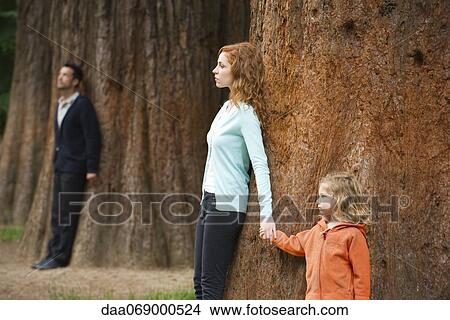 Picture - Mother and daughter leaning against tree, father standing separate in background. Fotosearch