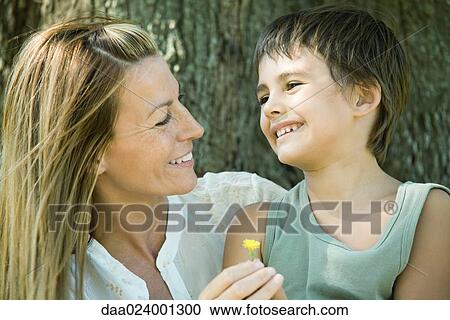Stock Image - Mother and son smiling at each other, woman holding flower, head and shoulders. Fotosearch