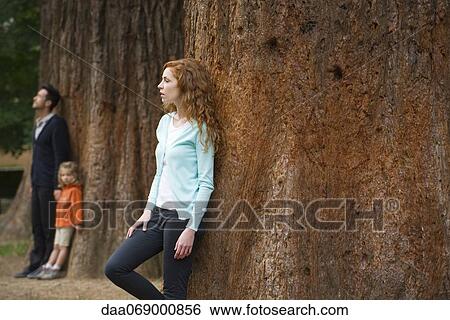 Mother leaning against tree trunk, husband and daughter separate in background View Large Photo Image Stock Photograph - Mother leaning against tree trunk, husband and daughter separate in background. Fotosearch
