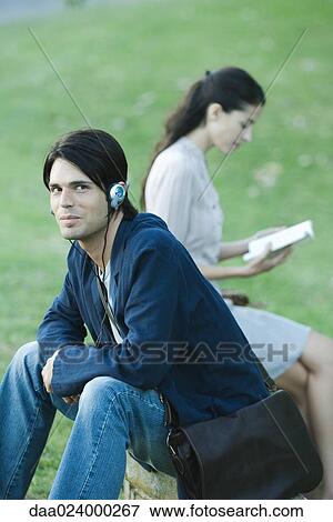 Stock Photo - People sitting in park, man listening to headphones while woman reads book. Fotosearch