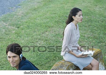 People sitting in park, man listening to headphones while woman reads book View Large Photo Image Stock Photography - People sitting in park, man listening to headphones while woman reads book. Fotosearch
