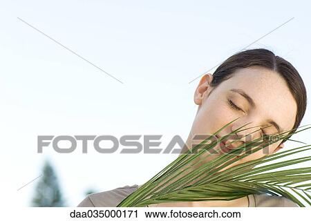 Stock Image - Portrait of young woman holding palm frond, eyes closed. Fotosearch