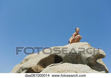 Picture - Preteen girl in bikini crouching on rock against blue sky, low angle view. Fotosearch