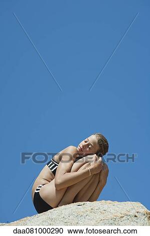 Stock Image - Preteen girl in bikini crouching on rock with eyes closed. Fotosearch