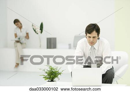 Stock Image - Professional man using laptop computer in casual office, female colleague reading in background. Fotosearch
