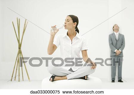 Stock Photography - Professional woman sitting on ottoman, drinking from water bottle, businessman and bamboo in background. Fotosearch