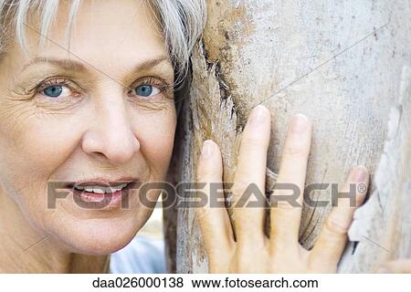 Senior woman leaning against tree trunk, smiling at camera, close-up View Large Photo Image Stock Photo - Senior woman leaning against tree trunk, smiling at camera, close-up. Fotosearch