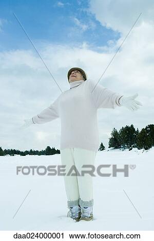 Stock Image - Senior woman standing on snow, arms out, looking up, low angle view. Fotosearch