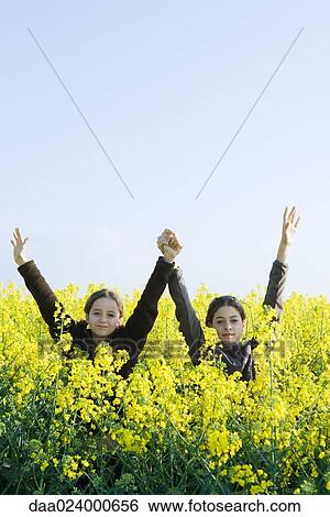 Stock Photograph - Sisters standing in field of canola in bloom, arms raised, looking at camera. Fotosearch