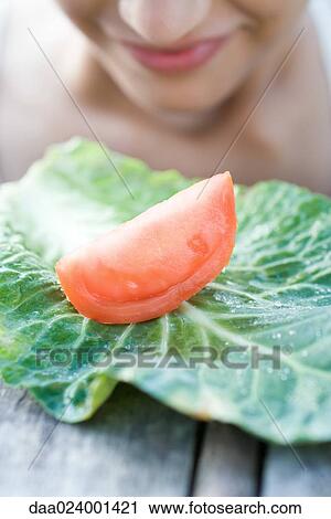 Stock Image - Slice of tomato on cabbage leaf, woman's face in background. Fotosearch