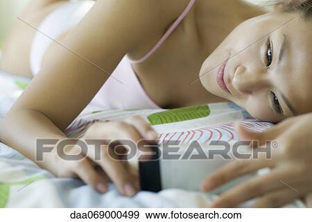 Smiling young woman lying on bed looking at cell phone View Large Photo Image Stock Photo - Smiling young woman lying on bed looking at cell phone. Fotosearch