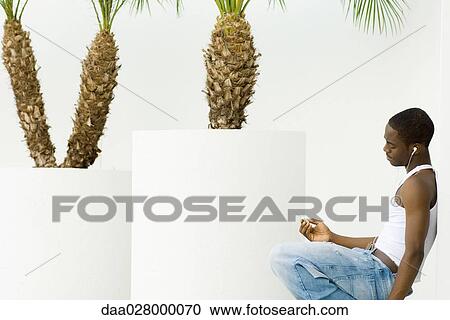 Stock Image - Teen boy listening to MP3 player, crouching, leaning against wall, potted palm trees in background. Fotosearch