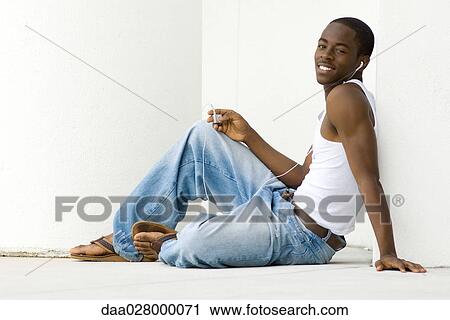 Stock Image - Teen boy sitting on floor, listening to MP3 player, smiling at camera, full length. Fotosearch