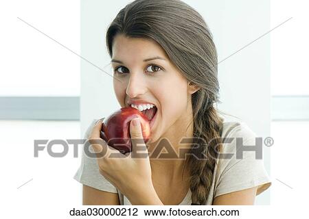 Stock Image - Teen girl biting into apple, looking at camera. Fotosearch