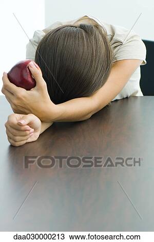 Stock Image - Teen girl holding apple, head resting on arms. Fotosearch