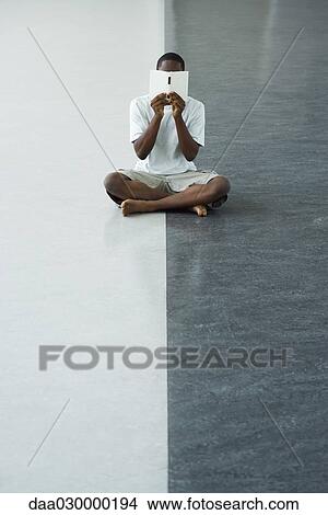 Teenage boy sitting on the ground, holding book in front of face View Large Photo Image Picture - Teenage boy sitting on the ground, holding book in front of face. Fotosearch