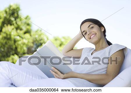 Picture - Teenage girl reclining in armchair, holding book, smiling at camera. Fotosearch