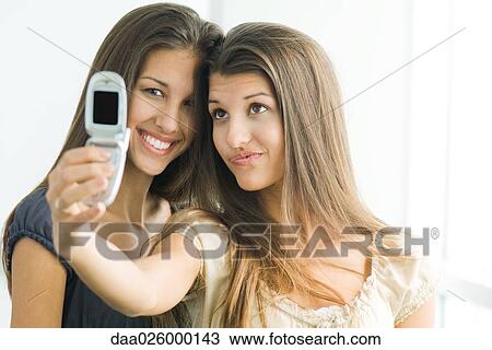 Stock Image - Teenage twin sisters photographing selves with cell phone, one smiling at camera. Fotosearch