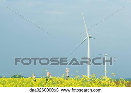 Stock Image - Two friends hiding in field of colza with arms raised, holding hands, wind turbines in background. Fotosearch