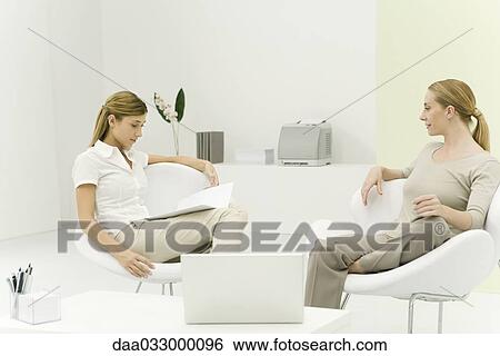 Two women sitting together in casual office, smiling, one reading document View Large Photo Image Stock Photograph - Two women sitting together in casual office, smiling, one reading document. Fotosearch
