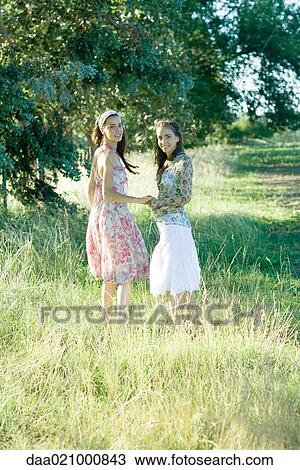 Stock Image - Two young women walking through field, holding hands. Fotosearch