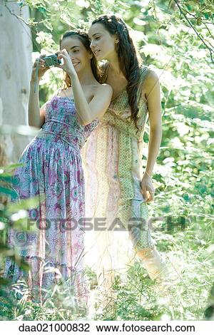 Stock Image - Two young women wearing sun dresses, standing in forest, looking at digital camera. Fotosearch