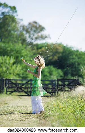 Stock Image - Woman blowing bubbles on rural path, side view. Fotosearch