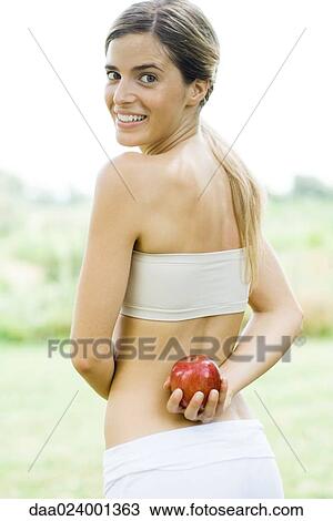 Woman holding apple behind back, looking over shoulder at camera View Large Photo Image Stock Image - Woman holding apple behind back, looking over shoulder at camera. Fotosearch