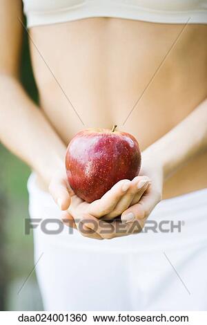 Stock Image - Woman holding apple in front of bare abdomen, close-up of mid section. Fotosearch
